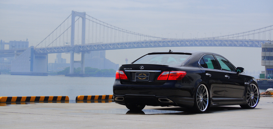 Black WALD Executive Line Lexus LS460L rear three-quarter view with a suspension bridge backdrop