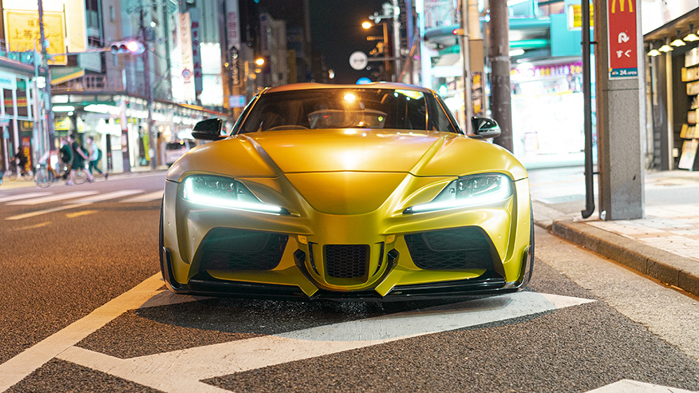 Yellow WALD wide body Toyota Supra head-on front view at night with illuminated headlights and front bumper spoiler