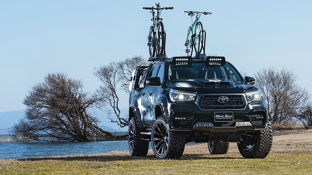 WALD Black Bison Toyota Hilux GUN125 with roof LED bar and bicycle carriers beside a lake