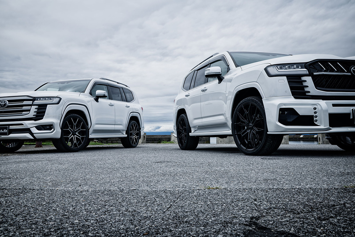 Two white Toyota Land Cruiser LC300 SUVs on WALD ELDORANTE E11C wheels — low side view by the water