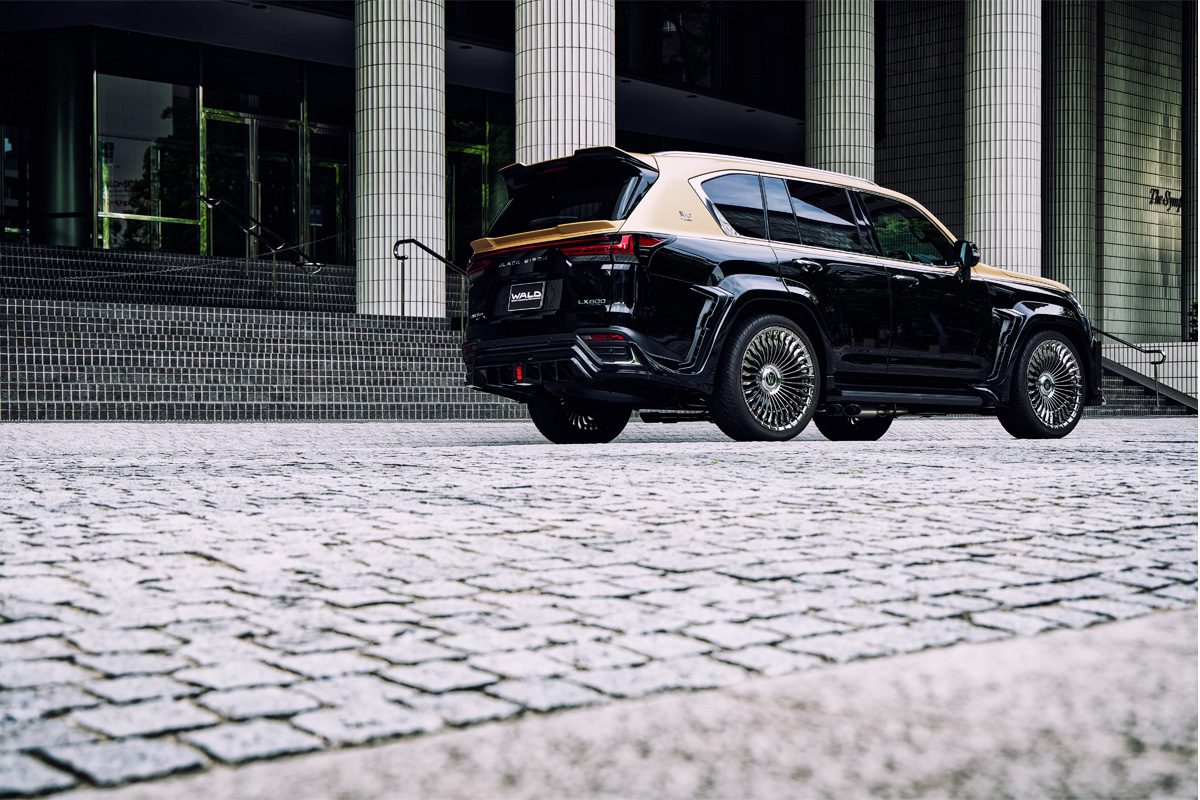 WALD Lexus LX600 Black Bison rear three-quarter on cobbles — rear skirt, tailgate spoiler and gold multi-spoke WALD wheels