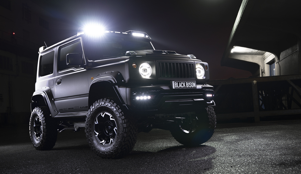 WALD Black Bison Edition Suzuki Jimny Sierra JB74W — matte black wide body Jimny front three-quarter night view with WALD front bumper spoiler, Japamericana grill and roof LED light bars