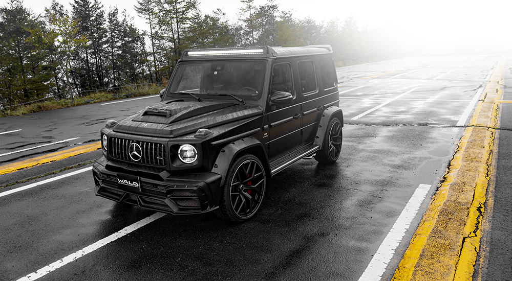 WALD Black Bison Mercedes G-Class W463A body kit — black G63 AMG with wide arch package, bonnet scoop and front roof spoiler LED bar on a wet mountain road