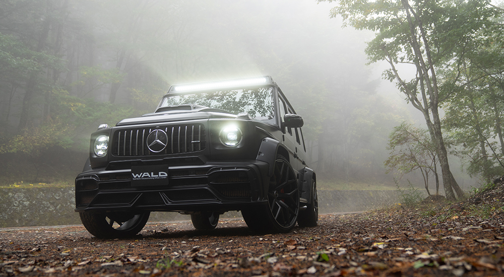 WALD Black Bison Mercedes-AMG G63 W463A front three-quarter in fog — black G63 with roof LED bar, bonnet scoop and wide arch package on forest road