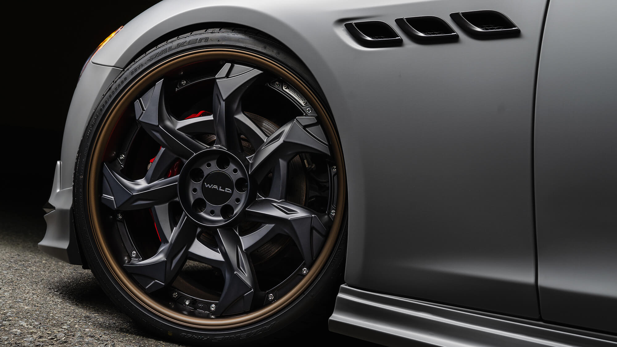 Close-up of a WALD Vorsalino V12C wheel in matte black with bronze alumite rim on a matte grey Maserati